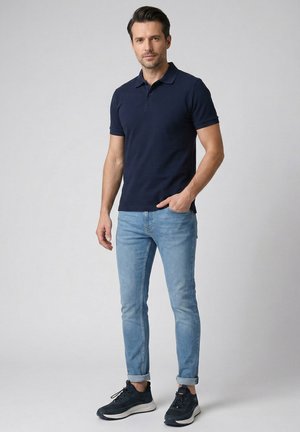 Man staat, draagt een marineblauw poloshirt, lichtblauwe opgerolde spijkerbroek en zwarte sneakers tegen een effen lichte achtergrond.
