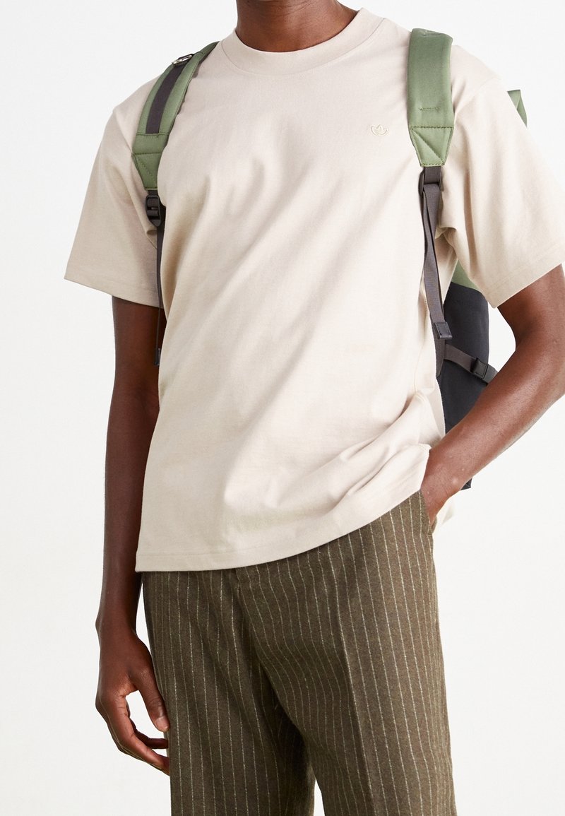 Mies, jolla on päällään beige t-paita, vihreät ja ruskeat raidalliset housut sekä musta ja vihreä reppu, käsi taskussa, neutraali tausta.