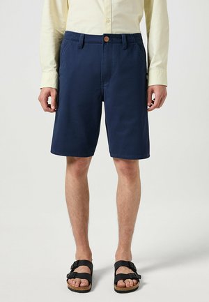 Marineblaue Shorts aus Baumwolle mit Knopfverschluss, seitlichen Taschen und einem lässigen Schnitt. Tragen mit schwarzen Sandalen.