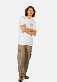 Weißes Baumwoll-T-Shirt mit einem schwarzen, runden Muster, kombiniert mit beigen Cargo-Hosen, die Seitentaschen haben. Getragen mit olivgrünen Sneakern.