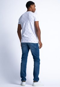 Tricou alb cu imprimeu mic albastru, combinat cu blugi albaștri. Modelul stă cu spatele spre cameră, purtând adidași albi.