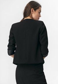 Kobieta z brązowymi włosami związanymi do tyłu, w czarnej marynarce z podwiniętymi rękawami i pasującą czarną spódnicą, stojąca tyłem na białym tle.