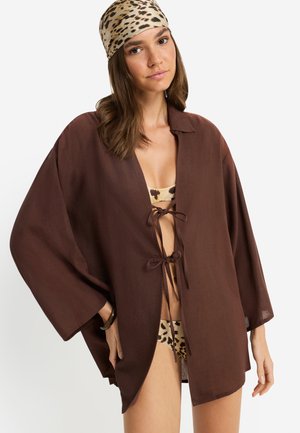Braunes, weit geschnittenes Wickelhemd mit Bindeband vorne und weiten Ärmeln. Darunter ein gemustertes Bikini in Beige- und Brauntönen.