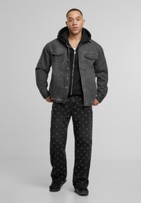 Vīrietis stāv ar rokām kabatās, ģērbies melnās sporta kurpēs, melnās biksēs ar baltām punktiem, melnā kapučjaka un pelēkā pogājama jakā.