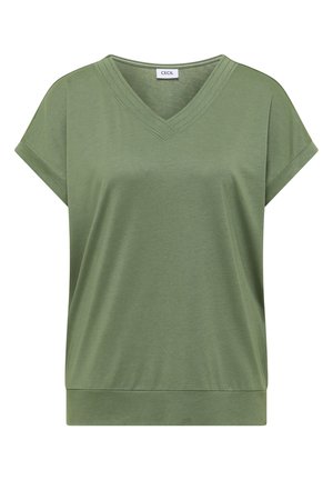 Femme aux cheveux courts marron portant un haut vert olive à manches courtes et un jean en denim bleu, vue de dos sur un fond uni.