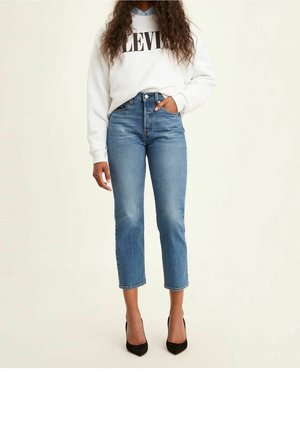 Femme portant un jean bleu court, un sweat-shirt blanc Levi’s, une chemise bleue à col en dessous et des talons hauts noirs pointus, debout.