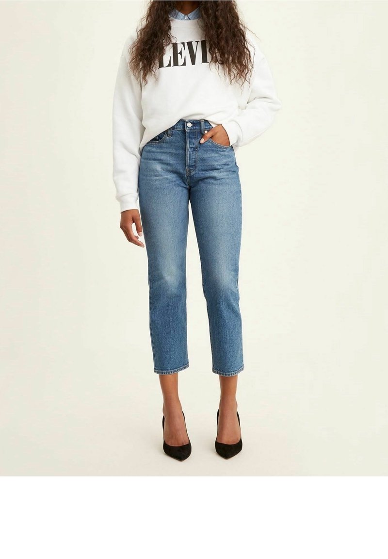 Femme portant un jean bleu court, un sweat-shirt blanc Levi’s, une chemise bleue à col en dessous et des talons hauts noirs pointus, debout.