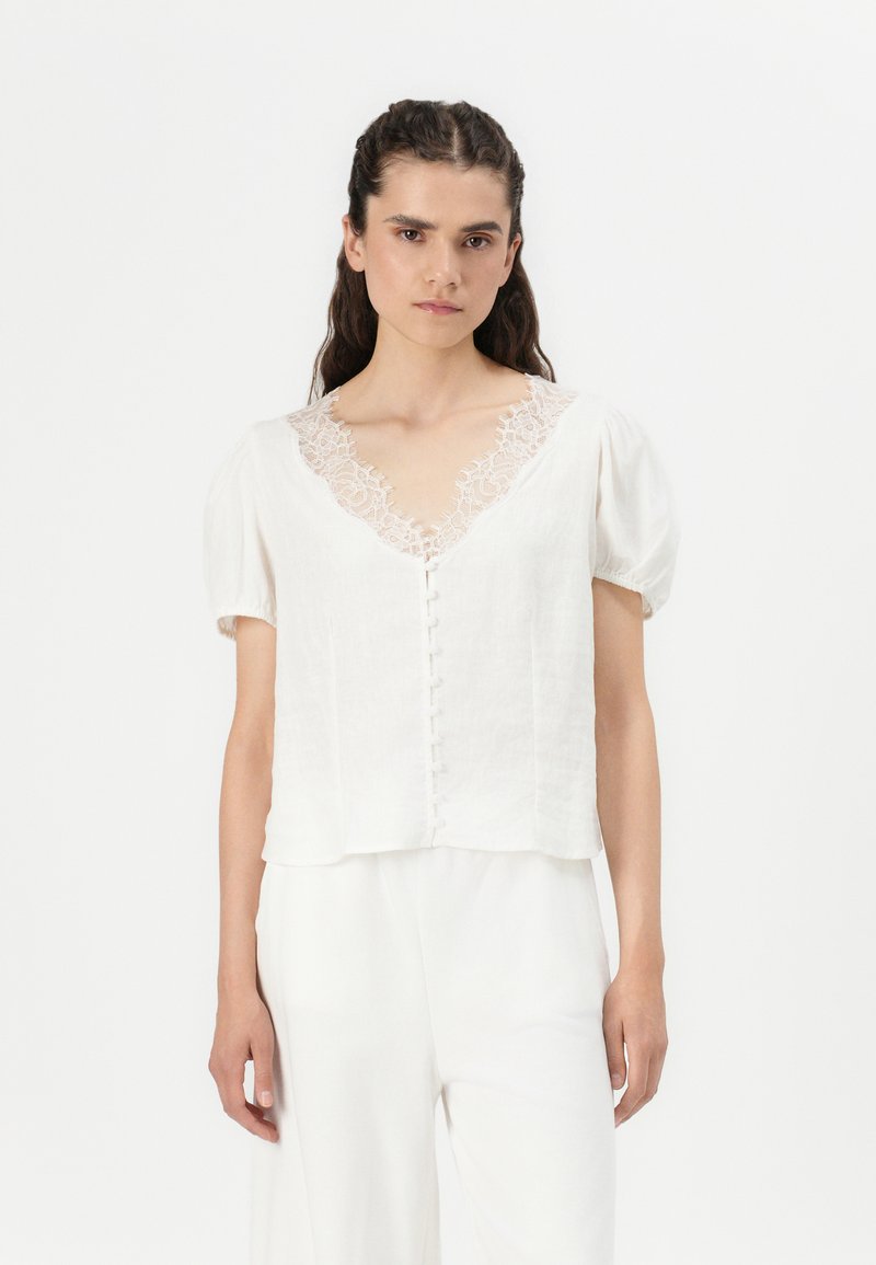 Blouse en lin blanc avec des manches courtes bouffantes, un décolleté en V agrémenté de dentelle, et un devant boutonné. Texture douce et coupe décontractée.