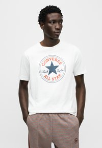 T-shirt en coton blanc avec un logo étoile bleu et un texte rouge et bleu, accompagné d'un pantalon à carreaux aux tons orange et bleu.