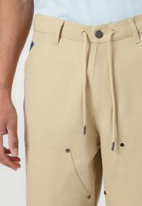 Beige cotton blend pants with a drawstring waistband, featuring stitched accents and two front pockets with snap closures.