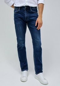 Vaqueros de mezclilla azul oscuro con un corte recto, con un ligero desgastado, cinco bolsillos y costuras en contraste. Combinados con zapatillas blancas.