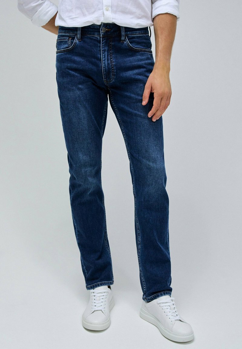 Vaqueros de mezclilla azul oscuro con un corte recto, con un ligero desgastado, cinco bolsillos y costuras en contraste. Combinados con zapatillas blancas.