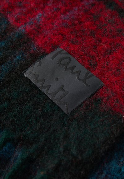 Textured wool fabric in dark green, red, and black with a black leather label featuring embossed text "Paul Smith." Notable pattern and color contrast.