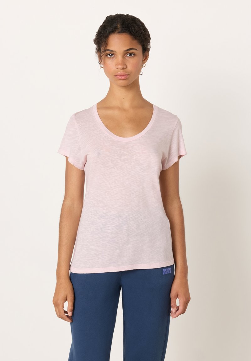 Svetlo roza majica s kratkimi rokavi in širokim okroglim izrezom, izdelana iz mehke, rahlo teksturirane tkanine, nošena z modrimi športnimi hlačami z logotipom blagovne znamke.