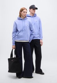 Hellviolette Kapuzenpullover mit Kordelzügen, dunkle Jeans mit weitem Bein und eine schwarze Tragetasche mit Markenlogo. Glatter Stoff und lockere Passform.