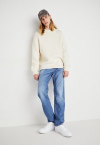 Crème-kleurige gebreide trui met geribbelde manchetten, blauwe denim jeans en witte sneakers. Het model draagt een grijze pet met een embleem.