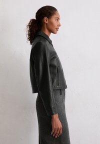 Czarna skórzana kurtka typu cropped z kołnierzem, kieszeniami bocznymi i ściągaczami na mankietach, noszona na szarej dzianinowej spódnicy. Gładka tekstura, dopasowany fason.