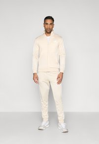 Αθλητική φόρμα σε ανοιχτό μπεζ χρώμα, κατασκευασμένη από λείο υλικό, με μπουφάν με φερμουάρ και όρθιο γιακά, καθώς και αντίστοιχο παντελόνι με στενή γραμμή.