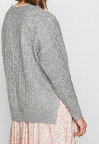 Grauer Kabelstrickpullover mit Rundhalsausschnitt und langen Ärmeln, ausgestattet mit seitlichen Schlitzen und gerippten Bündchen, darüber getragen über einem rosa Blumenkleid.