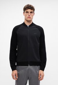 Svart zip-up jakke med ribbestrikket krage, kontrastfargede teksturerte ermer og sidelommer med glidelås. Har diskret merking på brystet.