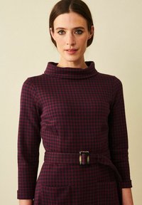 Sovittuva mekko viininpunaisena, jossa on houndstooth-kuvio. Vaatteessa on kaarroke kaulus, kolme neljäsosaa hihat ja vyötärön ympärillä vyö suoralla soljella.