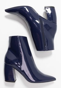 Botines de charol azul marino con punta afilada, cierre de cremallera y tacones de bloque, que presentan un acabado brillante y líneas marcadas.
