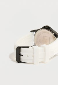 Ρολόι με μαύρη θήκη και λευκό λουράκι από σιλικόνη, με μαύρη πόρπη και κλείσιμο, φωτογραφημένο από πίσω σε λευκό φόντο.