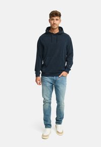 Mørkeblå hoodie med forlommer og snoretræk i hætten, parret med lyseblå jeans og hvide sneakers med beige detaljer.