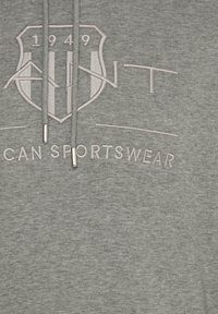 Gray hoodie featuring a textured finish, with a prominent shield logo and “1949” embroidered in light color. Metallic aglets on drawstrings.