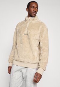 Béžový fleecový pulóver s vysokým golierom, polovičným zipsom a logom Champion na hrudi. Obsahuje zúžené manžety a lem pre tesné padnutie.
