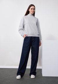 Sudadera gris con cuello alto, combinada con jeans oscuros de corte holgado. Zapatillas blancas completan el atuendo casual. Diseño simple y funcional.