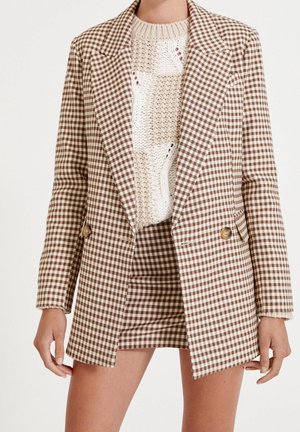 Kobieta w brązowo-białym blezerze w kratę vichy i dopasowanej spódnicy na kremowym, grubym dzianinowym swetrze, widoczna od szyi do połowy uda.