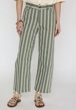 Donna che indossa pantaloni a gamba larga con strisce verticali verdi e bianche, sandali dorati e gioielli dorati alle mani.