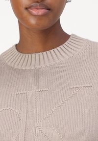 Maglione lavorato a maglia beige con un collo a costine e una lettera "K" in rilievo e ben visibile sul davanti. Tessuto liscio con un motivo sottile.