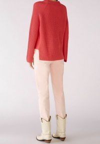 Personne portant un pull tricoté corail, un jean rose clair et des bottes de cowboy crème, se tenant dos à un fond uni.