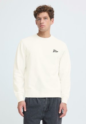 Felpa bianca con scollo rotondo con una piccola toppa triangolare nera con testo. Il tessuto sembra morbido con una texture liscia. Indossata con pantaloni grigio scuro.