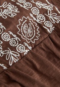 Tissu marron avec des motifs floraux brodés en blanc et deux motifs de poissons au centre, montrant des coutures texturées et un détail de couture froncée.