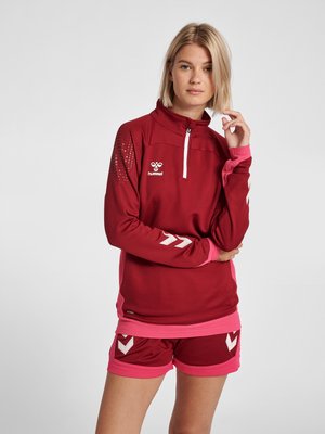 Lyshåret kvinde i mørkerød og lyserødt sportstøj med kvart-lynlås top og matchende shorts, stående med en hånd nær ansigtet og et neutralt udtryk.