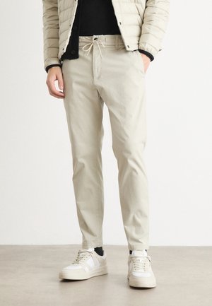 Pantaloni beige chiaro con vita regolabile e tasche laterali, abbinati a un giubbotto imbottito e sneakers bianche su un pavimento liscio.