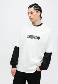 Weißes übergroßes T-Shirt mit schwarzem Print eines Keyboards und eines Herzens. Darunter ein schwarzes Langarmshirt getragen, kombiniert mit schwarzen Hosen.