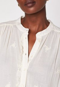 Primo piano di una persona che indossa una blusa bianco sporco con piccoli fiori ricamati, pieghe, patta dei bottoni arricciata e colletto alla mandarina.