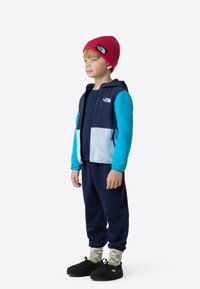 Kék és sötétkék polárdzseki cipzárral, világoskék panellel, fekete nadrág, zöld-fehér mintás zokni és piros kötött sapka.