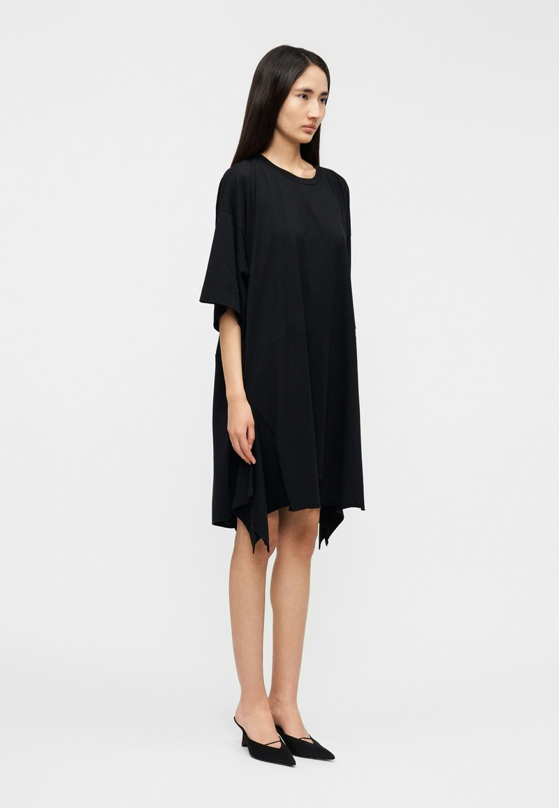 Robe noire surdimensionnée avec manches courtes, confectionnée dans un tissu doux. Présente une coupe décontractée et un ourlet asymétrique. Associée à des chaussures noires pointues.