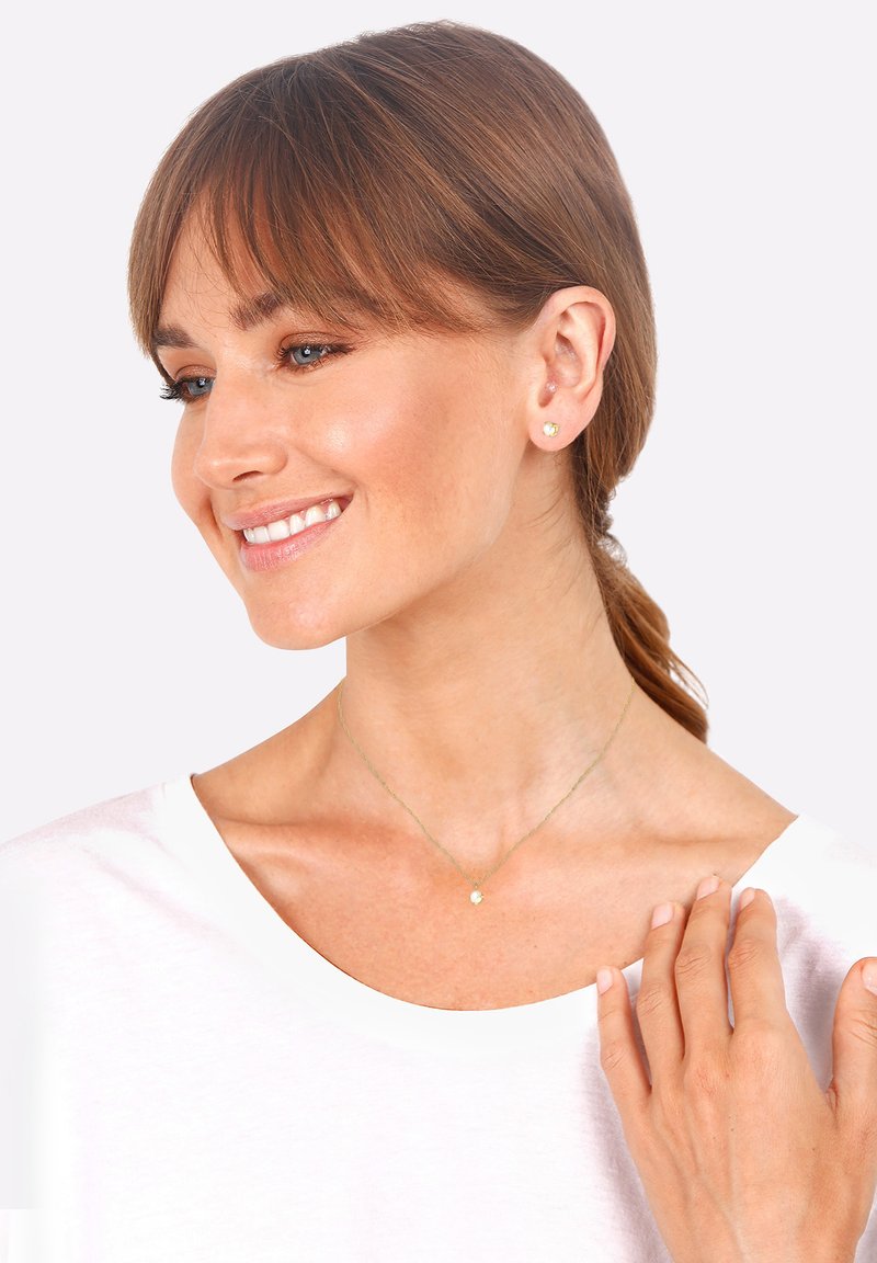 Orecchini a bottone in oro e una delicata collana d'oro con un piccolo ciondolo, indossati da una donna con capelli castano medio e una maglietta bianca.