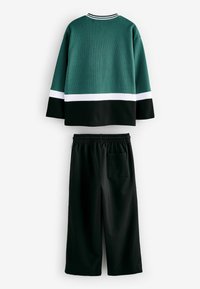 Grüner Kapuzenpullover mit weißem Streifen, kombiniert mit schwarzen weiten Hosen mit elastischem Bund und Gesäßtasche. Texturiertes Material über die gesamte Fläche.