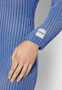 Main reposant sur la hanche portant un pull en grosse maille bleu avec une étiquette blanche indiquant "KOCHÉ" sur le bord de la manche.