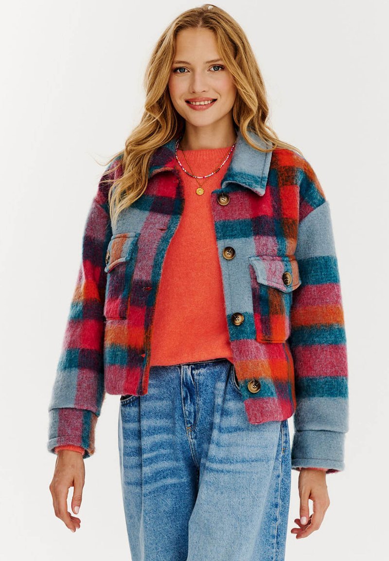 Pestrofarebný károvaný kabát v modrej, červenej a oranžovej vlne, s dvoma prednými vreckami a gombíkovými uzávermi, nosený cez oranžový sveter a džínsové nohavice.