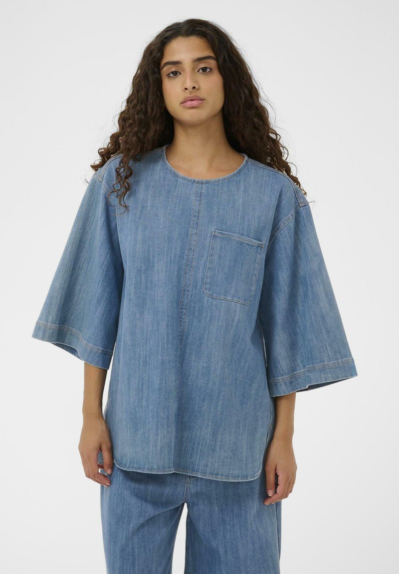 Haut en denim à coupe loose, col rond et manches courtes. Dispose d'une poche poitrine et de coutures visibles dans une teinte plus claire.