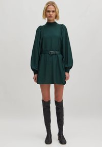 Dunkelgrünes Langarmkleid mit einem hohen Kragen und gerafften Schultern, tailliert mit einem passenden Gürtel, kombiniert mit schwarzen knielangen Stiefeln.