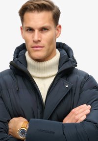Chaqueta de plumas azul marino con cuello alto y capucha, combinada con un suéter de cuello alto de canalé color crema. Presenta un logo y un reloj de pulsera con correa marrón.